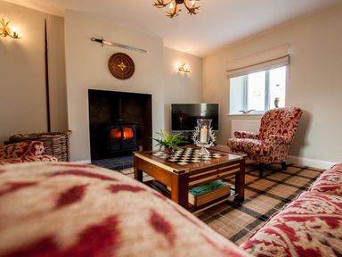 A living room with a fireplace a television patterned armchairs a chessboard coffee table and a window at Westmains Farm in Penton