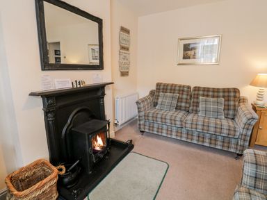 A living room with a fireplace stove a plaid sofa cushions a lamp and a wicker basket at Raglan Cottage in Bowness-on-Windermere