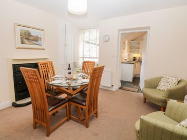 A dining room with a glass table and wooden chairs next to a fireplace and two green armchairs with cushions at Raglan Cottage in Bowness-on-Windermere