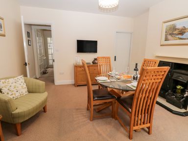 A dining area with a glass table and wooden chairs next to a fireplace and green armchair at Raglan Cottage in Bowness-on-Windermere