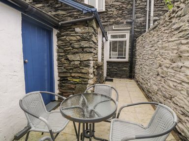 An outdoor patio area with a glass round table and three wicker chairs next to stone walls and a blue door at Raglan Cottage in Bowness-on-Windermere