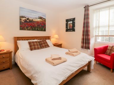 A bedroom with a wooden bed and white bedding two bedside tables with lamps a red armchair and a window with blinds and plaid curtain at Raglan Cottage in Bowness-on-WIndermere