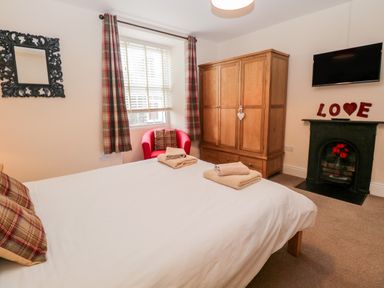 A bedroom with a bed towels wooden wardrobe armchair window with curtains mirror and wall-mounted television at Raglan Cottage in Bowness-on-Windermere