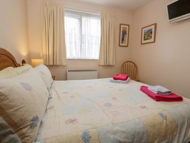 A bedroom with a bed covered in a floral patterned bedspread two framed pictures on the wall a wooden chair with folded towels and a wall-mounted TV at Treetops in Windermere
