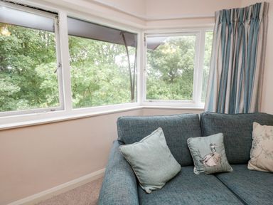 A corner sofa with three cushions near corner windows with blue and beige curtains at Treetops in Windermere