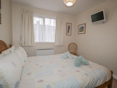 A bedroom with a bed pillows rolled towels a wooden chair two framed pictures a wall-mounted TV and a window with curtains at Treetops in Windermere