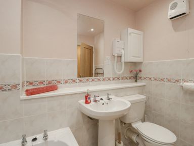 A bathroom with a sink a toilet a bathtub a mirror a wall-mounted hairdryer and a small cabinet at Treetops in Windermere