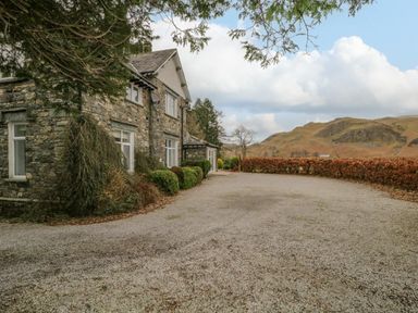 A stone house with bushes in front and a gravel driveway with hills in the background at The Old Vicarage in Keswick