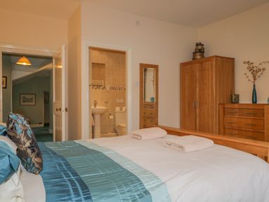A bedroom with a bed, wooden wardrobe, chest of drawers, mirror, and an en suite bathroom at The Old Vicarage in Keswick