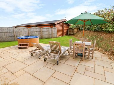An outdoor patio with two wooden lounge chairs a wooden table with four chairs and a green umbrella next to a hot tub and lawn at Ash Lodge in Greetham Rutland