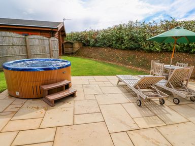 An outdoor patio with a hot tub wooden lounge chairs a table with chairs and a green umbrella at Ash Lodge in Greetham Rutland