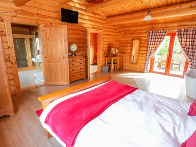 A wooden bedroom with a bed covered with white and red linens a dresser with a globe a wooden desk and chair plaid curtains on glass doors leading outside at Ash Lodge in Greetham Rutland
