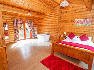 A bedroom with a wooden bed and red accents and a white bathtub near glass doors at Ash Lodge in Greetham Rutland