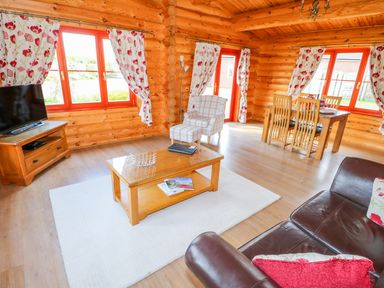 A living room with wooden walls red framed windows floral curtains a leather sofa armchair coffee table dining table and chairs at Ash Lodge Greetham Rutland