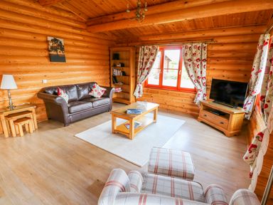A living room with a brown leather sofa, wooden coffee table, plaid armchair, wooden bookcase, and a television at Ash Lodge in Greetham Rutland