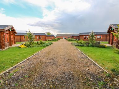 Gravel pathway flanked by green lawns and wooden cabins with orange window frames at Ash Lodge in Greetham Rutland