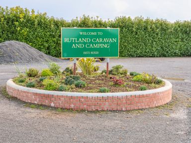A green welcome sign for Rutland Caravan and Camping on a circular brick flower bed by a gravel area with a hedge in the background at Ash Lodge in Greetham Rutland