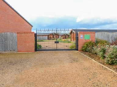 A gravel driveway leading to a black metal gate with brick pillars and a sign next to garden beds at Ash Lodge in Greetham Rutland