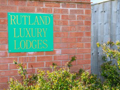 A green sign with yellow text reading Rutland Luxury Lodges on a brick wall next to a wooden fence with shrubs in front