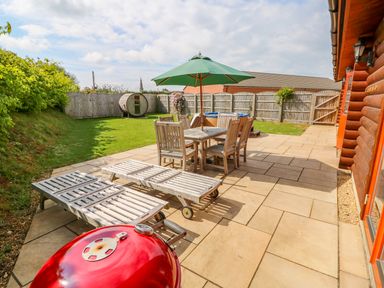 A patio with wooden chairs and table under a green umbrella two lounge chairs and a red barbecue in a fenced backyard at Yew Lodge in Greetham Rutland