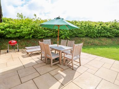 An outdoor patio with a wooden table and six chairs under a green umbrella and a red barbecue grill near some greenery at Yew Lodge in Greetham Rutland