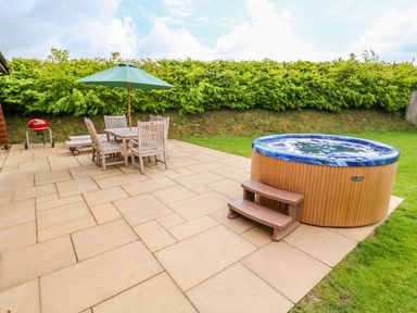 A patio with wooden table and chairs under a green umbrella next to a hot tub and a red barbecue at Yew Lodge in Greetham Rutland