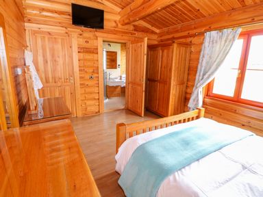 A bedroom with a wooden bed a blue blanket a wooden wardrobe a dresser a wall-mounted television an open door showing a bathroom and a window with curtains at Yew Lodge Greetham Rutland
