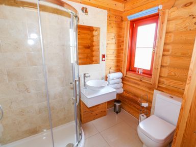 A bathroom with a curved glass shower enclosure a white sink with a faucet a stack of towels a window a toilet and wooden log walls at Yew Lodge in Greetham Rutland