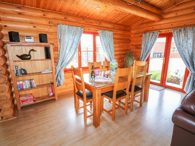 A dining area with wooden table and chairs next to windows with blue curtains and a wooden shelf with books and board games at Yew Lodge in Greetham Rutland