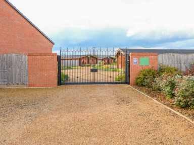 A gated entrance with a black metal gate between brick pillars and gravel driveway with wooden cabins behind at Yew Lodge in Greetham Rutland