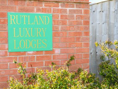 A green sign with yellow text reading Rutland Luxury Lodges mounted on a brick wall with green bushes and a wooden fence nearby at Yew Lodge in Greetham Rutland