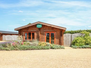 A wooden cabin with orange window frames and a green sign reading Yew surrounded by plants and gravel in front at Yew Lodge in Greetham Rutland