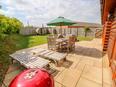 An outdoor patio with wooden sun loungers a dining table with six chairs and an umbrella a red barbecue grill and a fenced grassy yard at Willow Lodge in Greetham Rutland