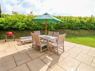 An outdoor patio with wooden chairs, a table with a green umbrella, two lounge chairs, and a red barbecue grill at Willow Lodge in Greetham Rutland