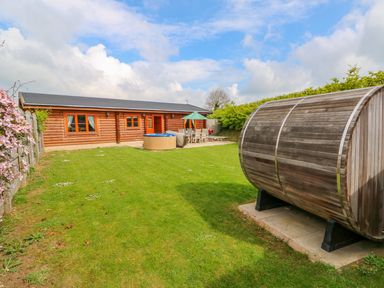 A wooden cabin with a patio hot tub table and chairs and a wooden barrel sauna in a garden at Willow Lodge in Greetham Rutland