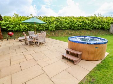 A patio area with wooden table and chairs under a green umbrella hot tub and a red barbecue grill at Willow Lodge in Greetham Rutland
