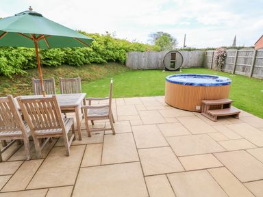 A backyard patio with wooden table and chairs under green umbrella a hot tub and a round wooden sauna at Willow Lodge in Greetham Rutland