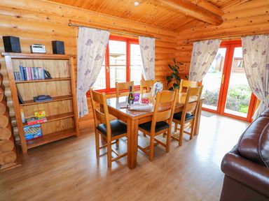 A dining area with a wooden table and six chairs near windows and glass doors with floral curtains in a log cabin at Willow Lodge in Greetham Rutland