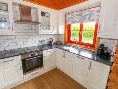A kitchen with white cabinets black countertops an oven a stovetop a kettle a coffee machine and a window with floral blinds at Willow Lodge in Greetham Rutland