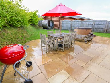 An outdoor patio with a wooden table and chairs under a red umbrella a red barbecue a wooden bench a hot tub a wooden shed and a fence at Sycamore Lodge in Greetham Rutland