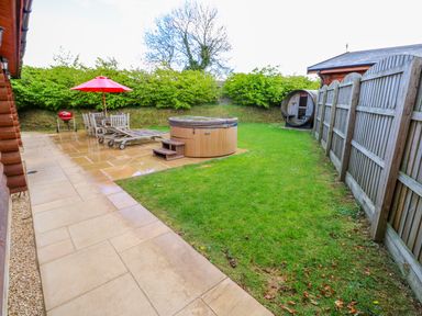 A backyard with a hot tub wooden lounge chairs a table with umbrella and a barbecue grill at Sycamore Lodge in Greetham Rutland