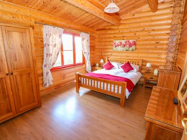 A bedroom with a wooden bed with red pillows and throw a wooden wardrobe and a wooden dresser with a mirror in a log cabin at Sycamore Lodge in Greetham Rutland