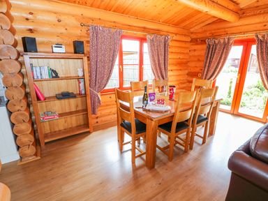 A wooden dining table with six chairs next to windows with curtains and a wooden bookshelf in a log cabin at Sycamore Lodge in Greetham Rutland