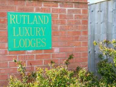 A green sign with yellow text reading Rutland Luxury Lodges mounted on a red brick wall with shrubs and a wooden fence nearby