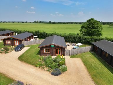 Three wooden lodges with fenced patios and parked car on gravel drive with green fields and trees at Sycamore Lodge in Greetham Rutland