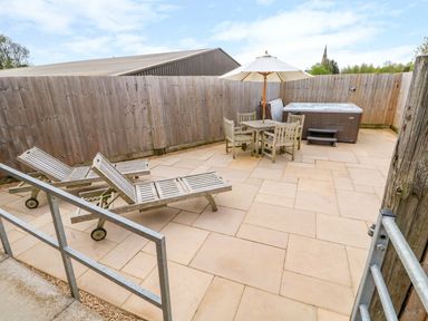 A patio area with two wooden sun loungers a wooden table with four chairs and an umbrella and a hot tub at Church View in Greetham Rutland
