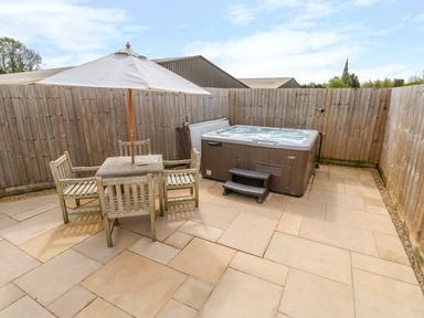 A patio area with wooden table and four chairs under a white umbrella next to a hot tub and wooden fence at Church View in Greetham Rutland