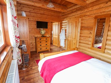 A bedroom with wooden walls a bed with red blanket a chest of drawers a wall mirror and a standing fan at Church View in Greetham Rutland