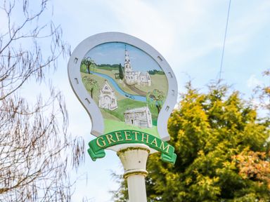 A village sign with illustrations of buildings and a river with trees in the background at Greetham in Rutland