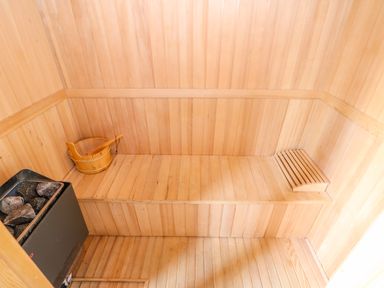 A wooden sauna room with a bench a wooden bucket and ladle and a heater with stones at Cedar Lodge in Greetham Rutland